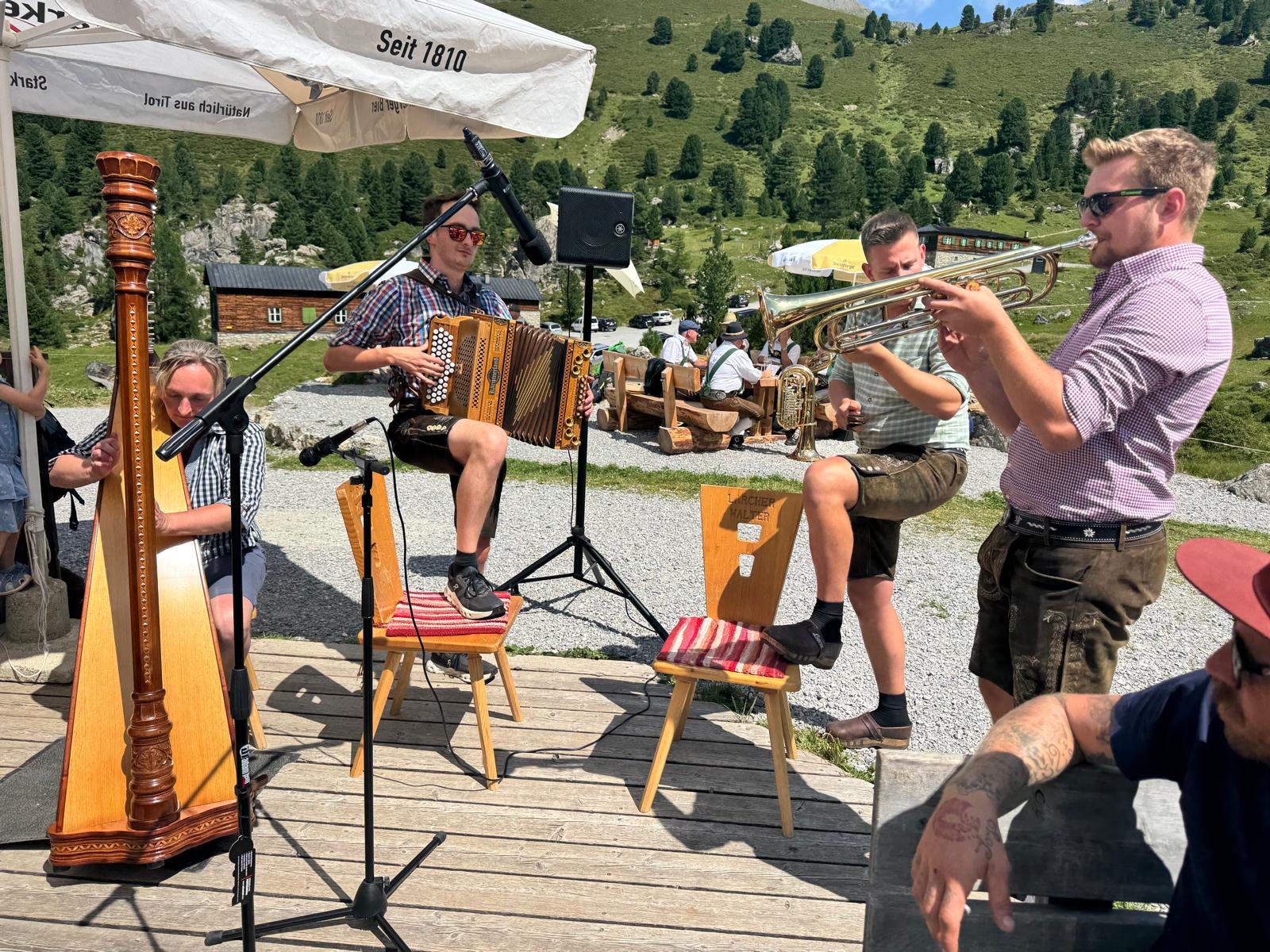 Junge Musikanten in Tracht mit Harfe, Steirischer Harmonika und Flügelhorn spielen auf der Holzterrasse der Lizumer Hütte.