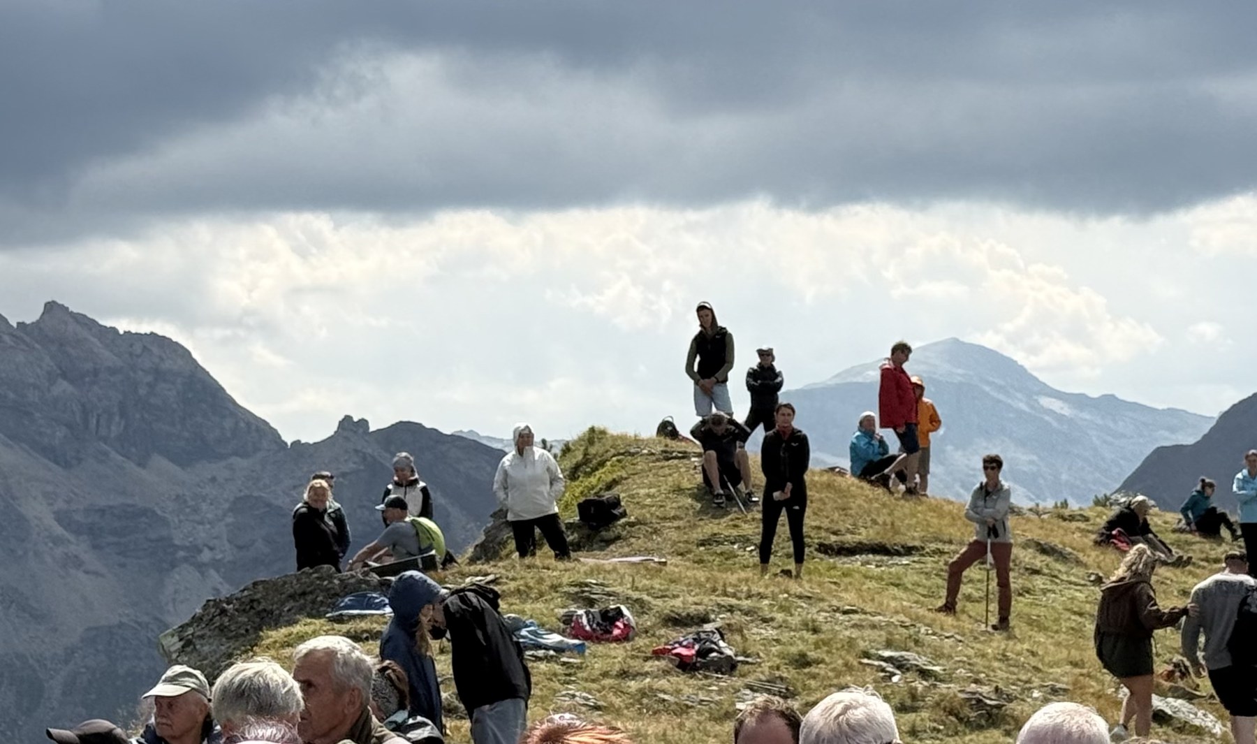 Zahlreiche Besucherinnen und Besucher sitzen und stehen am Berggrat, dahinter Wolken und Bergpanorama.