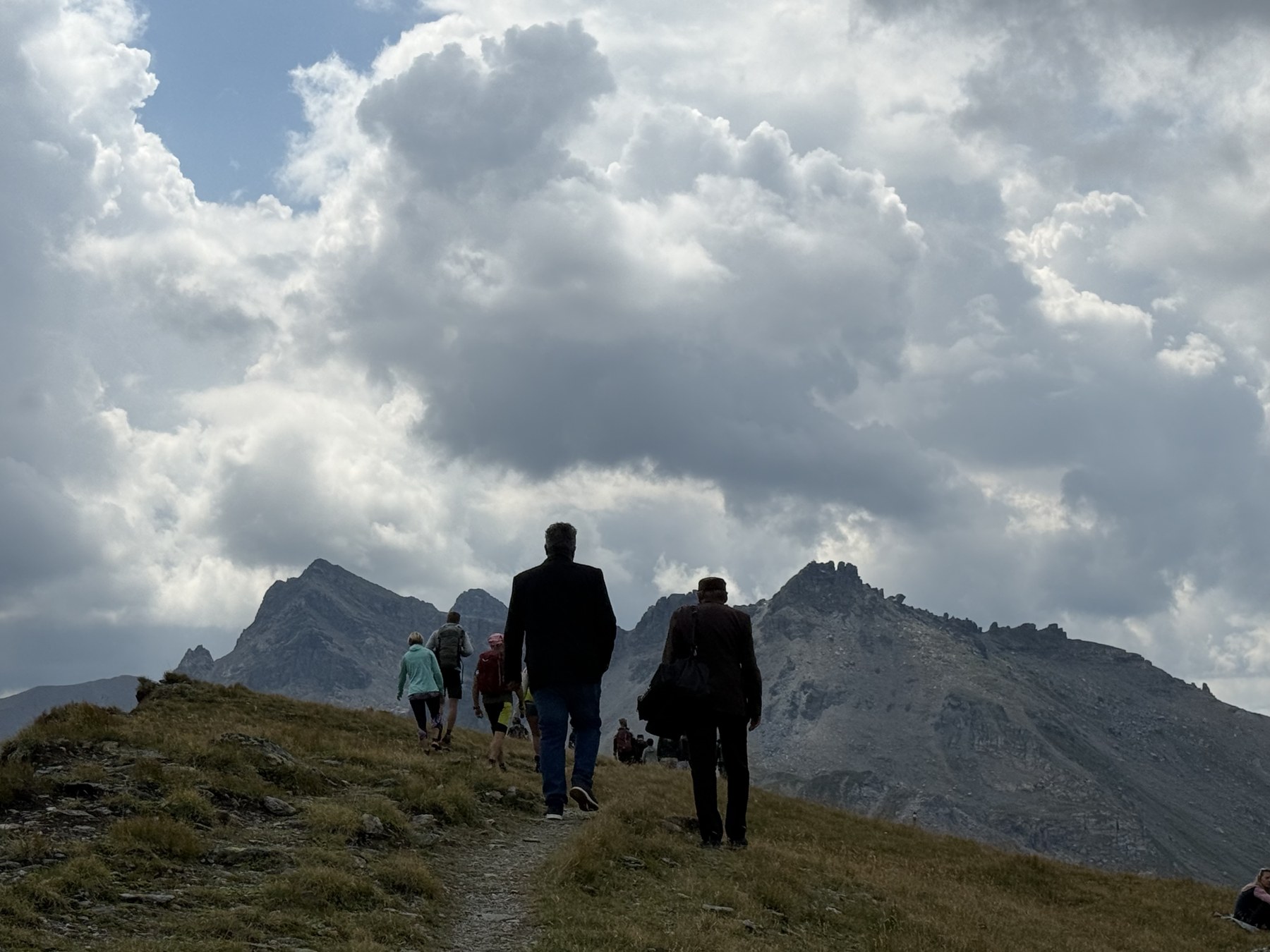 Besucherinnen und Besucher steigen über den Grat zur Mölsbergmesse auf, im Hintergrund Wolkenstimmung und Felsgipfel.