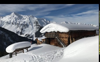 Tief verschneite Almhütte mit meterhohen Schneewechten auf dem Dach am Lann Niederleger, dahinter schneebedeckte Berggipfel unter blauem Winterhimmel im Wattental.