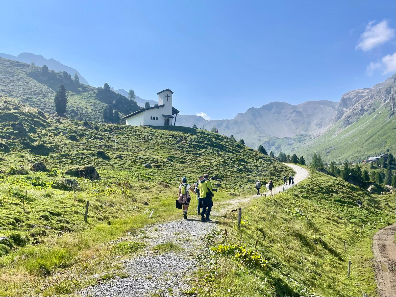 Besucherinnen und Besucher wandern auf dem Schotterweg zum Soldatenkirchlein in der Lizum bei strahlendem Sonnenschein.