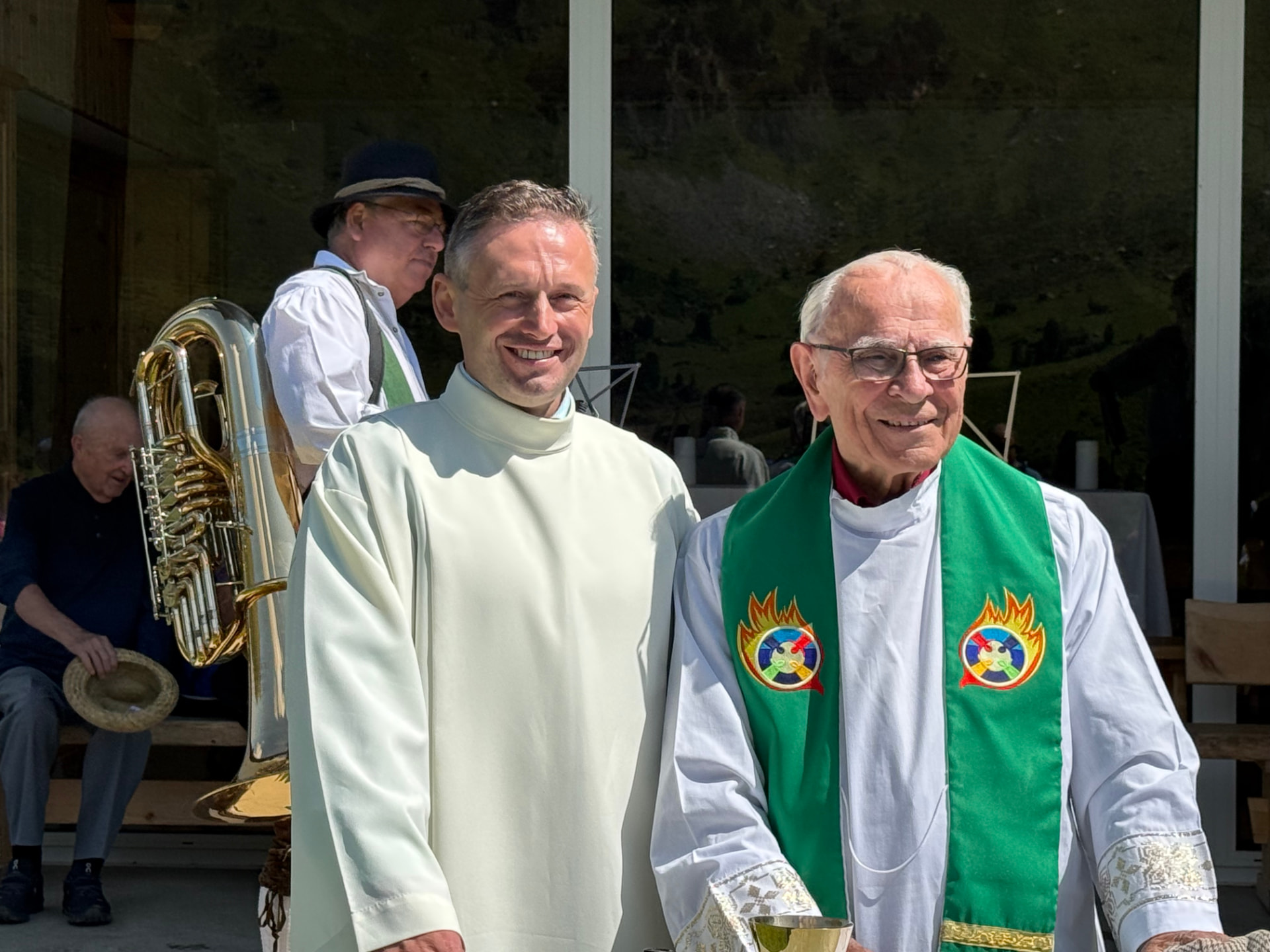 Zwei Priester in liturgischen Gewändern nach der Almerermesse, im Hintergrund ein Musikant mit Tuba.
