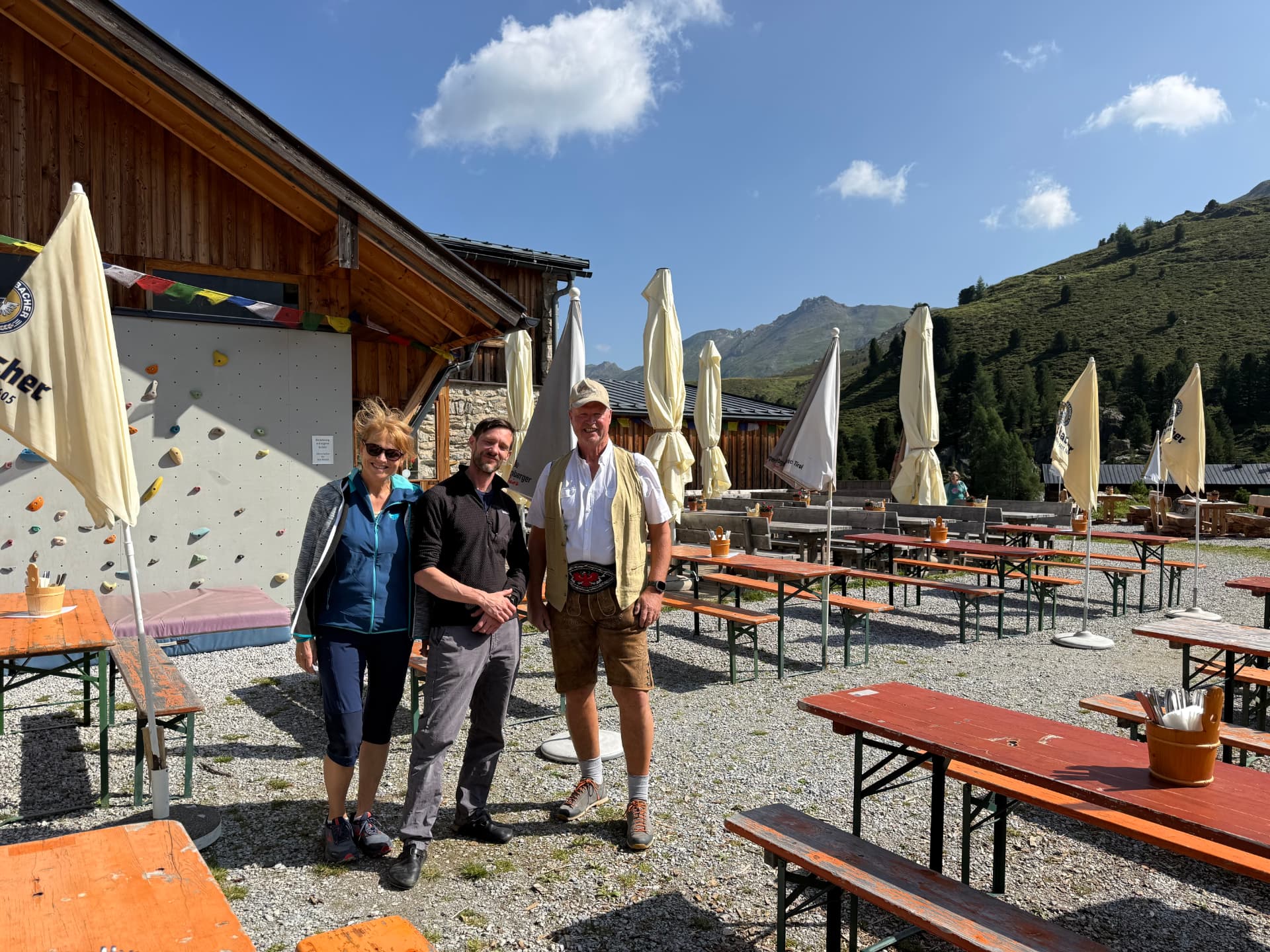 Schriftführerin, Hüttenwirt und Obmann stehen vor der Lizumer Hütte bei strahlendem Sonnenschein und blauem Himmel.