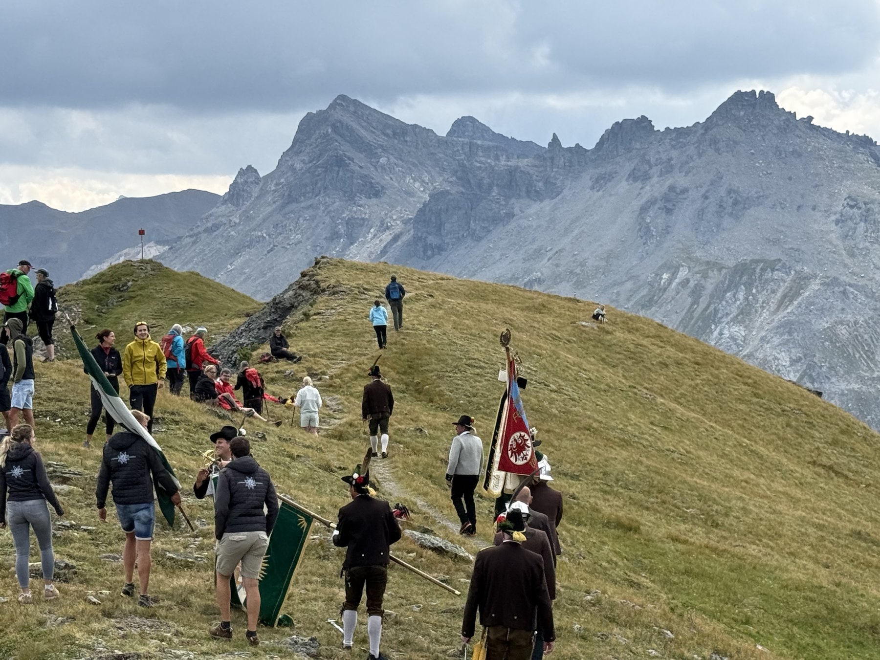 Prozession mit Fahnen und Musikanten zieht über den Bergrücken, dahinter schroffe Felsgipfel.