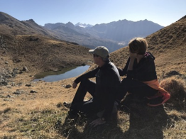 Zwei Personen sitzen auf einem grasigen Hang und blicken auf einen kleinen Bergsee und die schroffen Gipfel des Wattentals – weitläufiger Panorama-Ausblick.