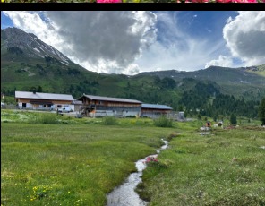 Almgebäude auf grüner Wiese, ein klarer Wiesenbach mäandert im Vordergrund durch den Lizumer Talboden, dahinter Berge unter dramatischem Wolkenhimmel.