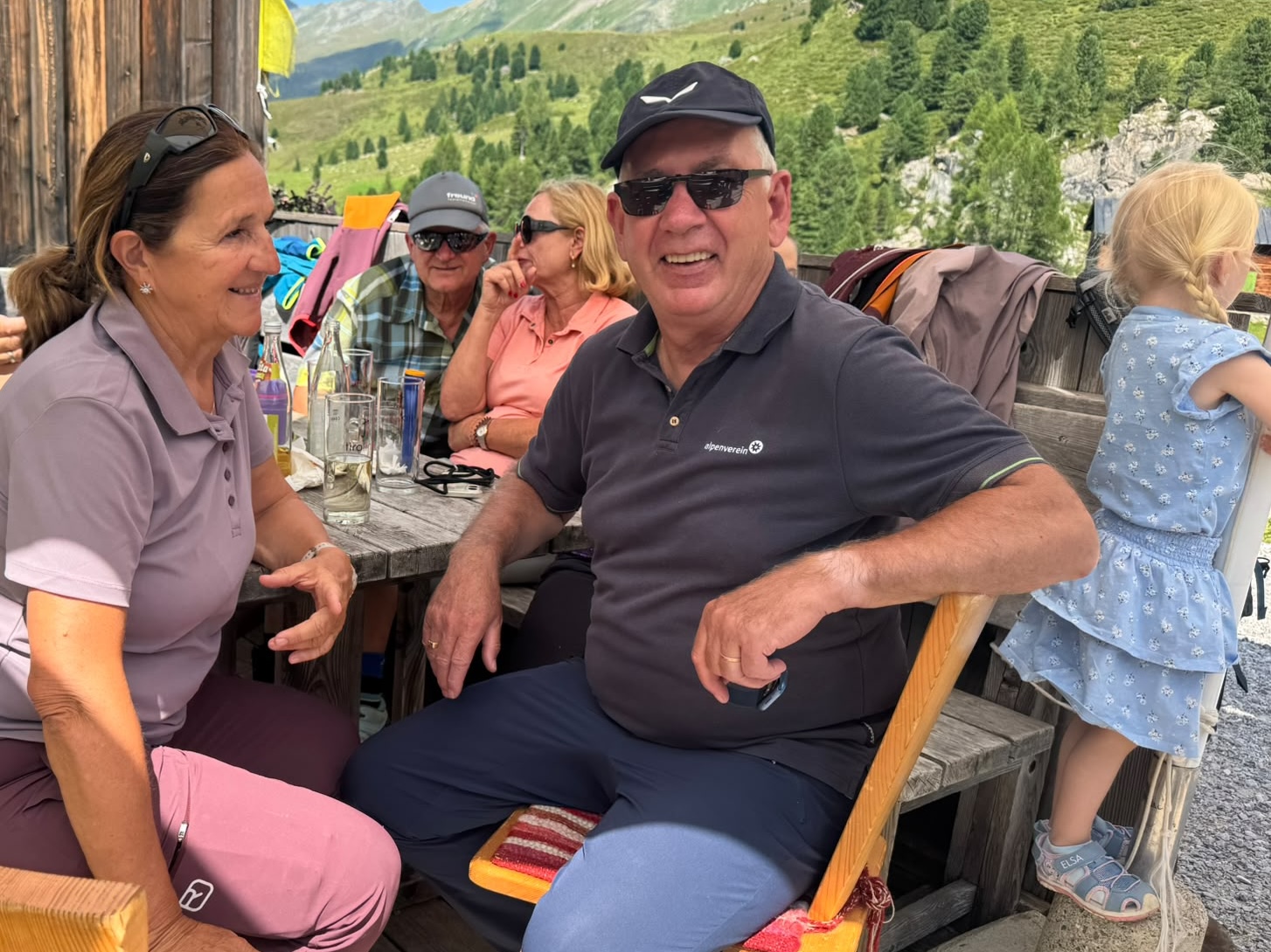 Gäste sitzen lachend an einem Holztisch vor der Hütte, im Hintergrund grüne Berghänge.