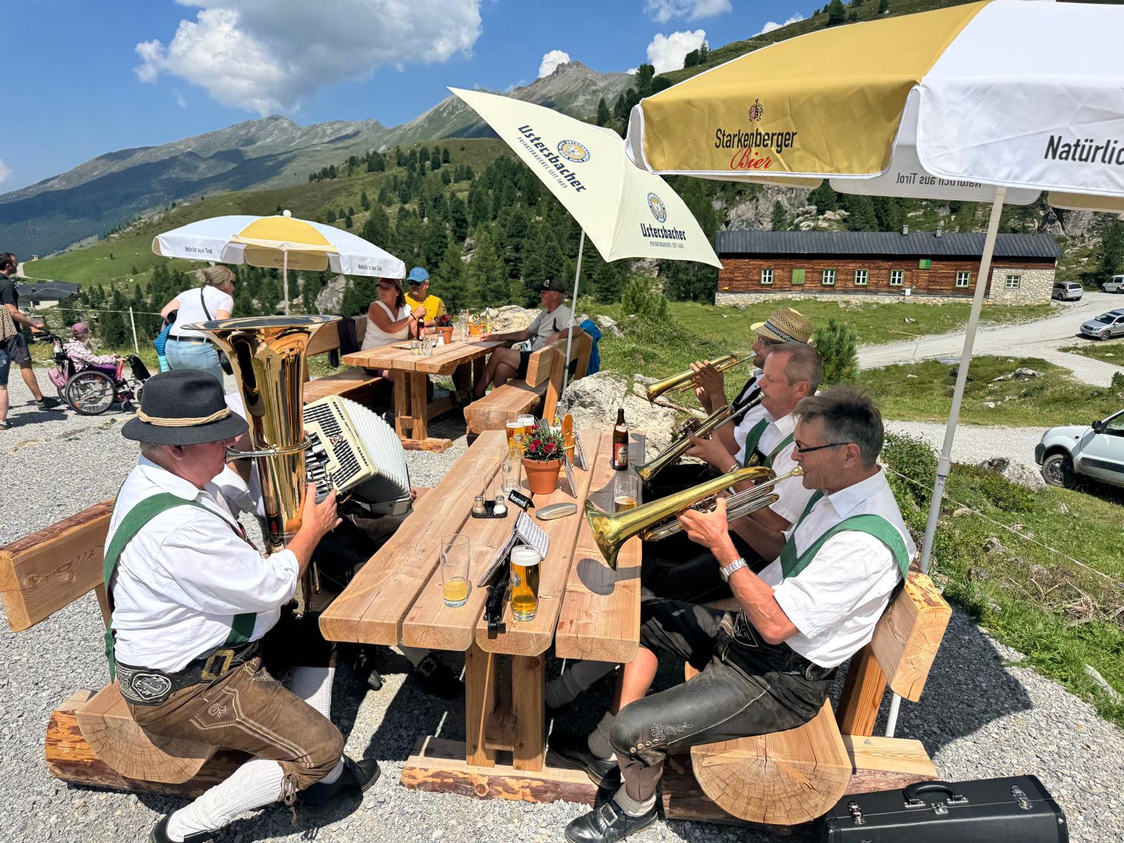 Musikanten in Tracht mit Tuba, Steirischer Harmonika und Flügelhörnern spielen an einem Holztisch vor der Lizumer Hütte.